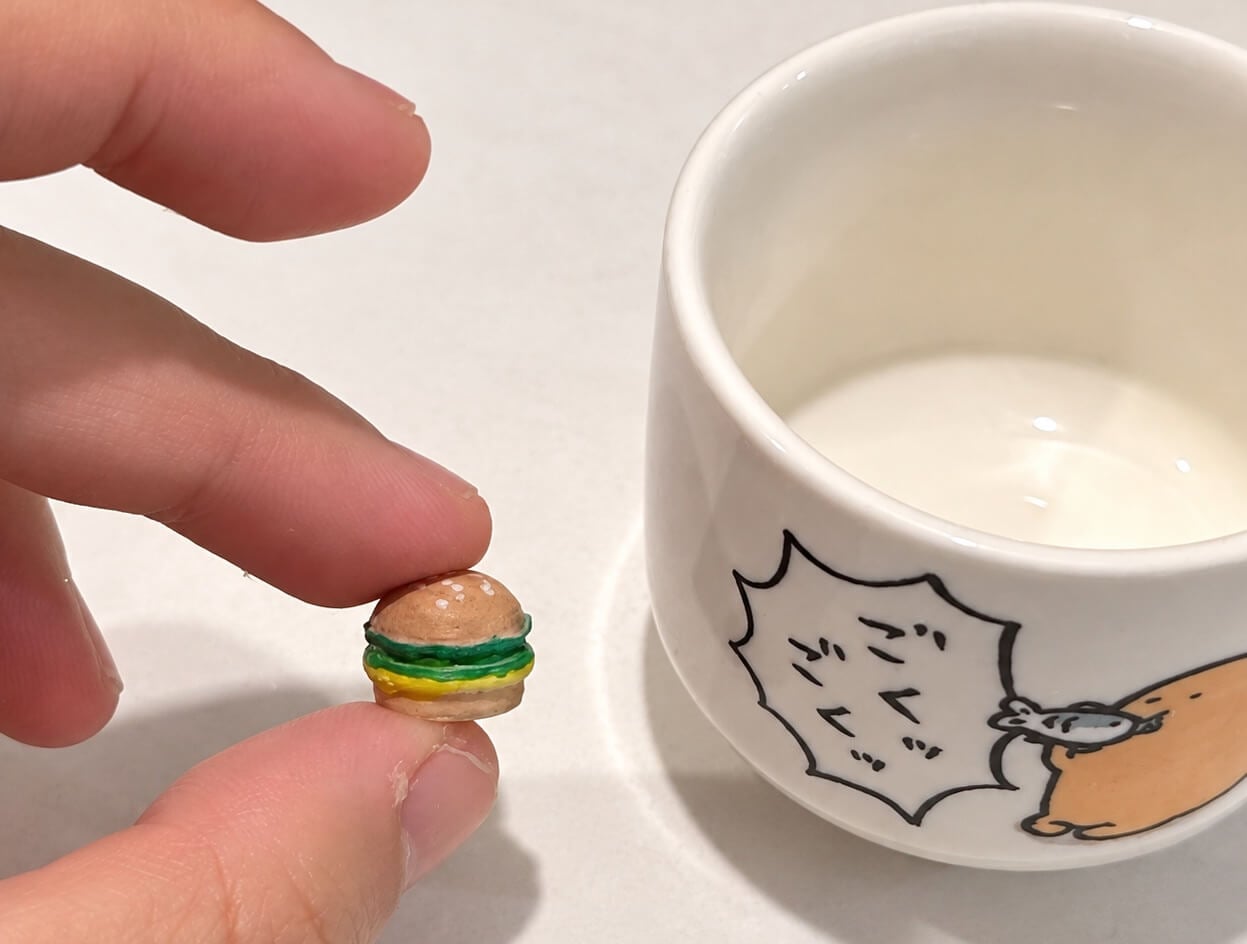Hand-painted miniature burger held between fingertips beside a ceramic cup for scale.